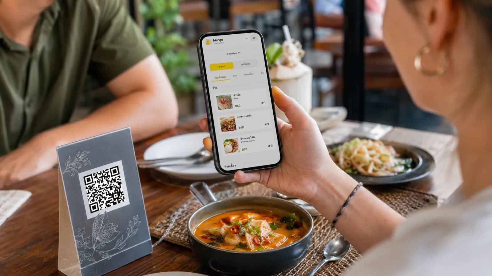 นักท่องเที่ยวที่โต๊ะอาหารกำลังสแกนเมนู QR หลายภาษาบนโทรศัพท์